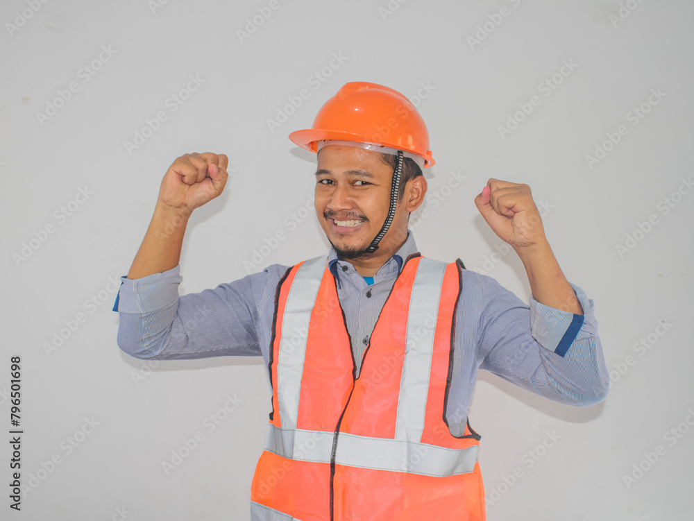 Asian man worker wearing safety helmet lookis happy celebrating his victory by clenching his fists against grey background
