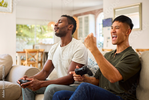 Same Sex Male Couple Or Friends Sitting On Sofa At Home Playing Computer Game Together