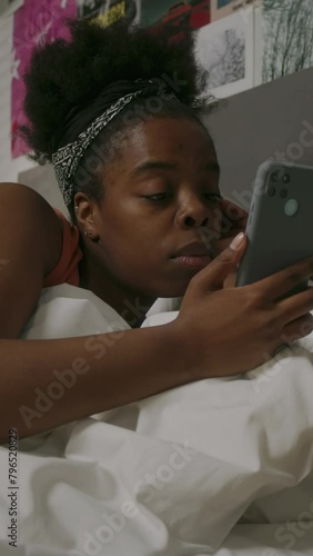 Vertical of young sleepy African American woman picking up cellphone from bedside table and checking up time while sleeping on soft bed in cozy bedroom with bright posters on wall
