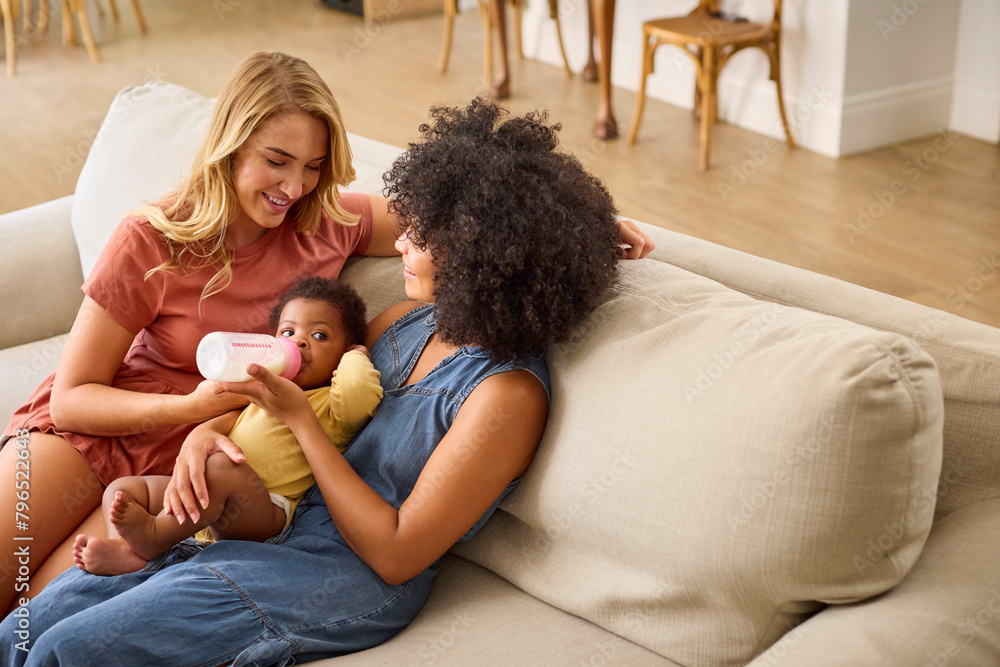 Same Sex Female Couple Or Friends Feeding Baby Girl With Bottle Sitting On Sofa At Home Together Same Sex Female Couple Or Friends Feeding Baby Girl With Bottle Sitting On Sofa At Home Together