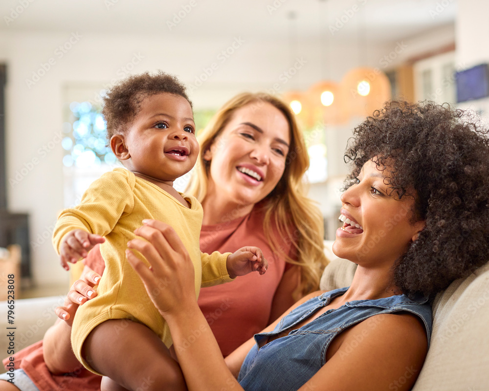 Same Sex Female Couple Or Friends Playing With Baby Sitting On Sofa At Home Together