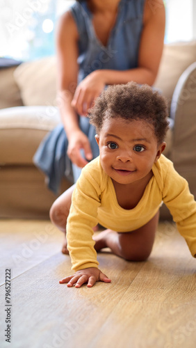 Smiling Baby Girl Learning To Crawl On Floor At Home With Mother Encouraging In Background photo