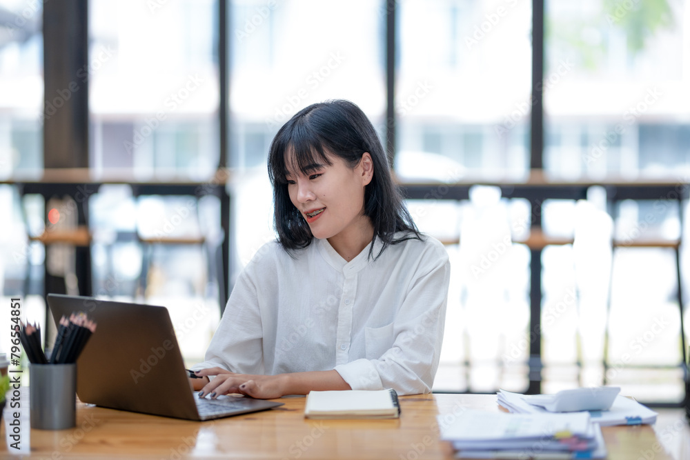 Cheerful young businesswoman with a pleasant smile working on her laptop at a bright office desk, with notebooks and pens nearby. Cheerful young businesswoman with a pleasant smile working on her laptop at a bright office desk, with notebooks and pens nearby.