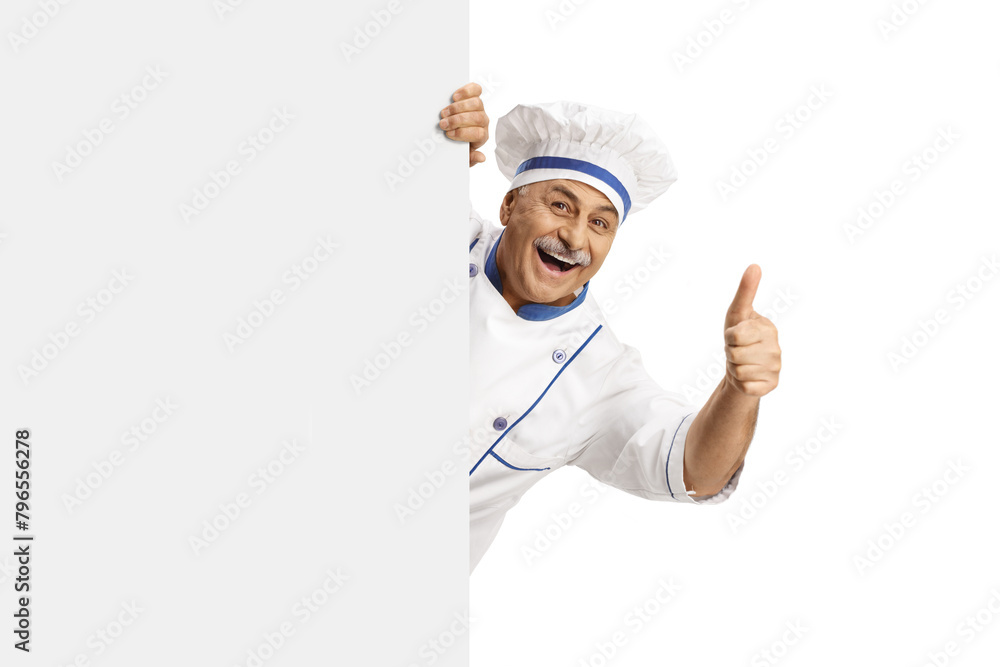 Mature male chef in a uniform gesturing thumbs up behind a blank panel Mature male chef in a uniform gesturing thumbs up behind a blank panel