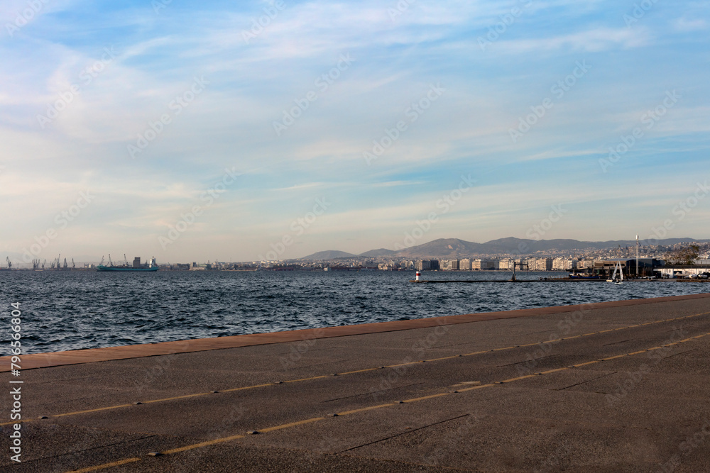 A scenic coastal pathway in Thessaloniki, Greece offers expansive views of the glistening sea with a city skyline and mountains in the distance.
