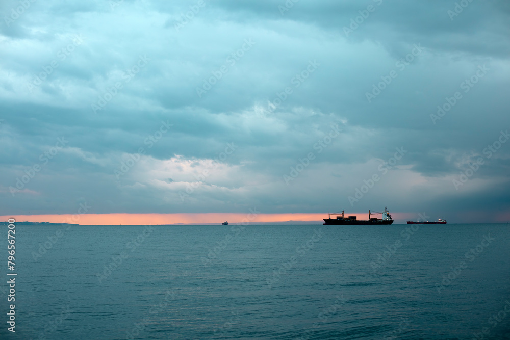 A solitary cargo ship sails across the calm sea as the sunset paints a pink hue on the horizon.
