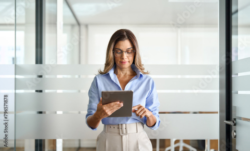 Busy middle aged businesswoman manager wearing eyeglasses looking at tablet standing in office. Mature professional business woman executive using tab, female entrepreneur or hr working with pa.