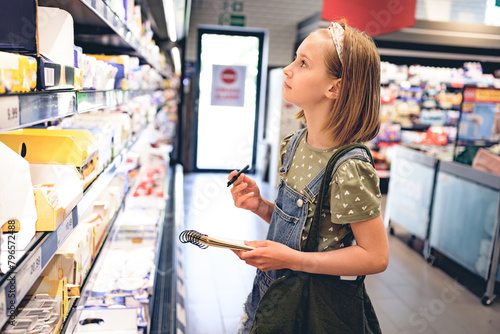Pretty girl child buying with shopping list in supermarket and looking productson shelf. Beautiful female preteen kid in grocery store photo