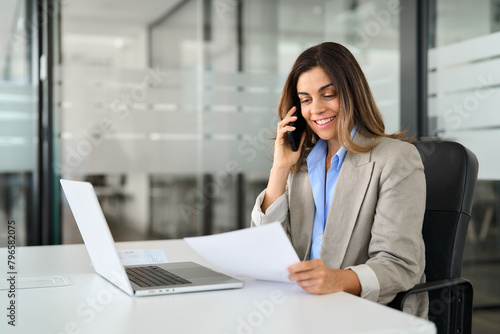Happy middle aged professional business woman executive making call having conversation at work. Mature female manager or entrepreneur talking on the phone checking document sitting at office desk.