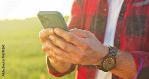 Cropped view of the optimistic caucasian farmer scientist in plaid shirt using smartphone mobile technology application while working at the field and smiling. Agriculture concept