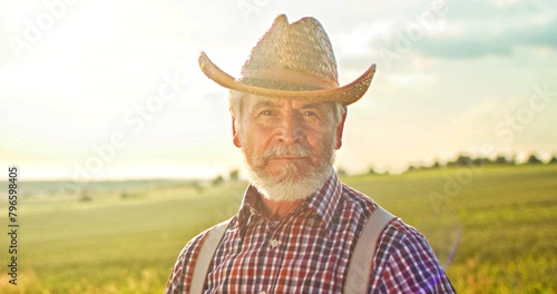 Portrait shot of senior caucasian man in hat standing in green field, smiling cheerfully to camera and feeling joy. Male farmer with smile outdoors in summer