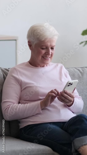 Old cheerful and friendly Caucasian woman using phone during weekend leisure time a home. Engaged in online shopping or reading news. Vertical video.