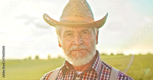 Portrait of senior smile man with beard looking to the camera in the golden field on the blue sky background sunny day. Face of the happy farmer worker at the summer nature. Farming people concept