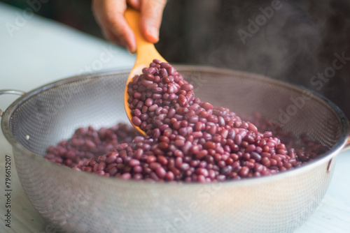 Woman's hand release temperature by using wooden ladle scoop boiled azuki beans in stainless steel sieve before mixed with tempeh starter, isolated on white table, closed up.