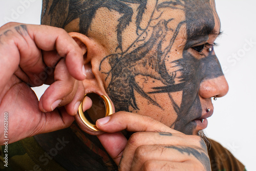 An asian man with extensive face tattoos fixes or puts on his flesh tunnel or large plug into his earlobes. photo