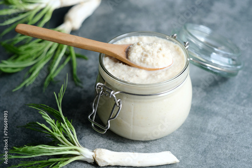 Handmade horseradish sauce in a jar with a spoon on top, horseradish roots on a gray background. Healthy vegan food, natural ingredients photo