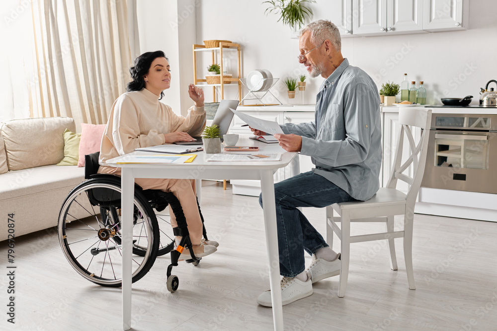 A man sits at a table next to a woman in a wheelchair in their kitchen at home. A man sits at a table next to a woman in a wheelchair in their kitchen at home.
