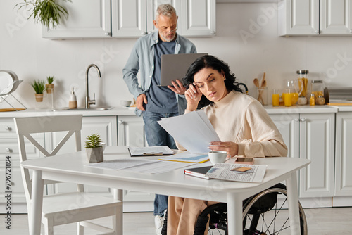 A disabled woman in a wheelchair gazes at a piece of paper, her husband by her side, in their kitchen at home.