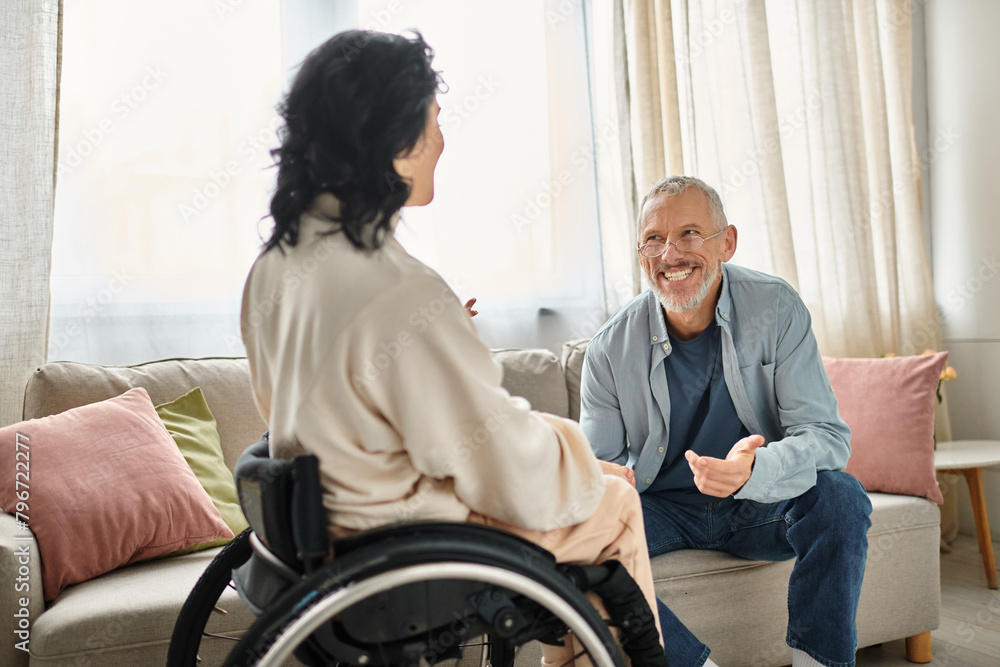 A woman in a wheelchair engages in conversation with her husband in their cozy living room setting. A woman in a wheelchair engages in conversation with her husband in their cozy living room setting.