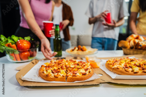 .Young Asian students gather with friends for a pizza party, laughing and sharing slices. Enjoying fast food delivery, they embody diversity and togetherness in a relaxed, enjoyable lifestyle.