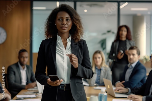A woman in a business suit stands in front of a group of people, holding a cell phone. Concept of professionalism and authority, as the woman is likely a leader or presenter in a meeting or conference
