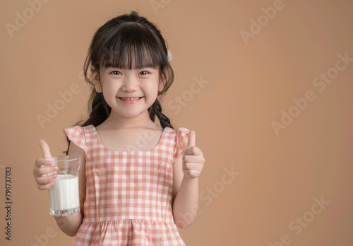Portrait of happy asian girl child holding milk glass and showing thumbs up, wearing pink gingham dress isolated on beige background with copy space