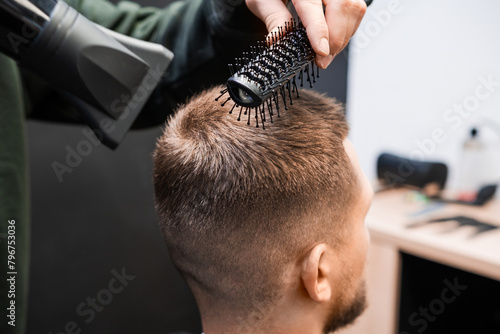 Stylist brushes man short hair using blowing dryer in barbershop closeup. Skilled barber does elegant hairstyle in beauty salon. Male haircut