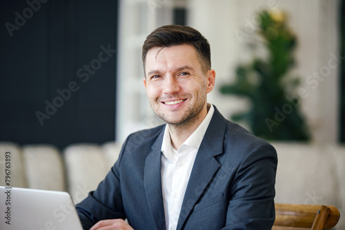 A contented businessman in a tailored suit shares a warm smile, laptop in tow, in a well-appointed home office.