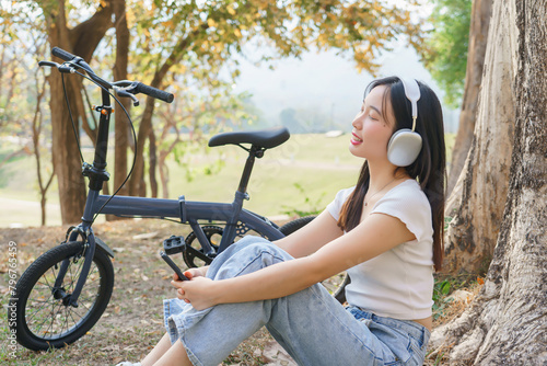 Young asian women wearing headphone and using smartphone to listening audio music with happiness while sitting on the grass to relaxation with journey travel lifestyle after cycle bicycle in the park