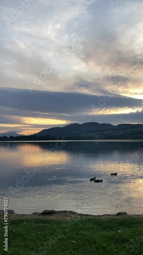 A tranquil evening scene of ducks swimming in a calm lake with the sun setting over the mountains in the background. The cloudscape is very atmospheric - Lake of Menteith, Stirling, Scotland photo