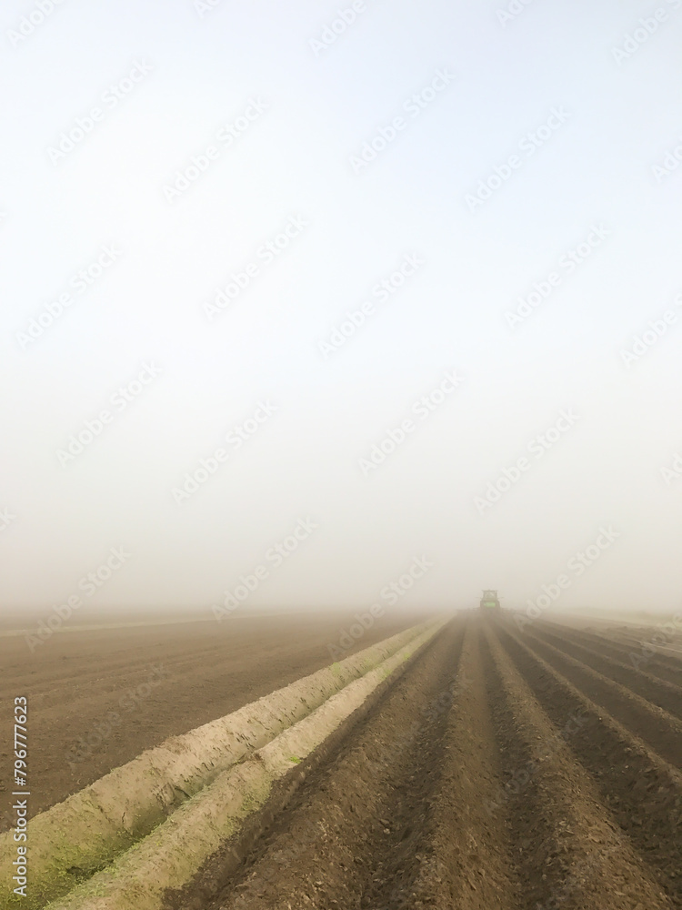Agricultural field with water furrows being tilled by a tractor driving into the fog