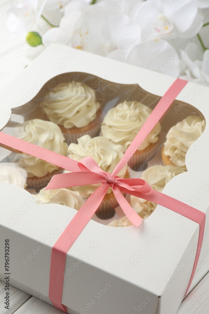 Tasty cupcakes with vanilla cream in box and orchid flowers on white wooden table, closeup Tasty cupcakes with vanilla cream in box and orchid flowers on white wooden table, closeup