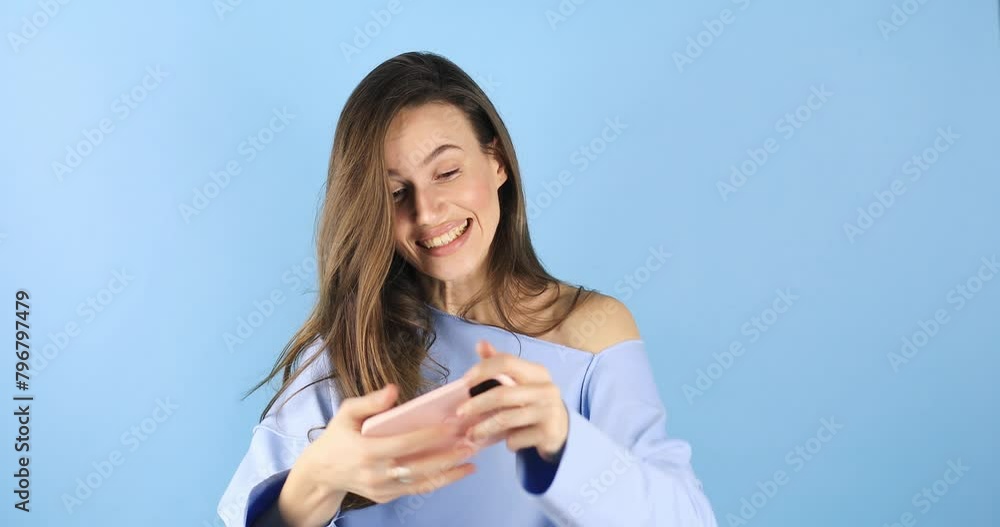 Brunette woman using play mobile cell phone hold gadget smartphone video pc games isolated on blue background. Happy woman play in video game on mobile phone. Brunette woman using play mobile cell phone hold gadget smartphone video pc games isolated on blue background. Happy woman play in video game on mobile phone.