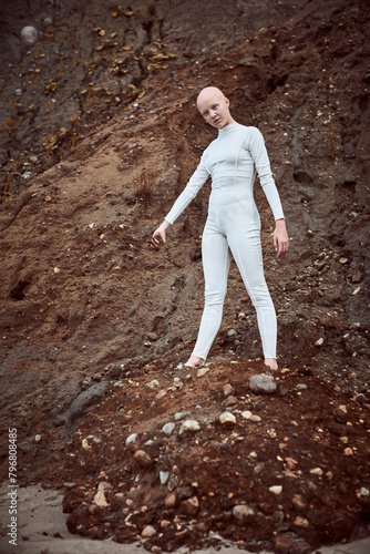 Full length portrait of young hairless girl with alopecia in white futuristic suit holding handful of barren soil thoughtfully  highlighting potential for coexistence between humanity and environment