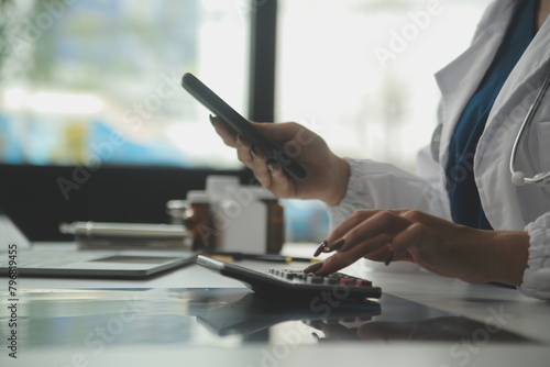 Serious female doctor using laptop and writing notes in medical journal sitting at desk. Young woman professional medic physician wearing white coat and stethoscope working on computer at workplace.