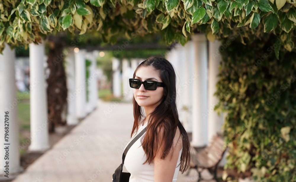 Portrait of a young woman wearing sunglasses, in the park with a beautiful sunset light. The white pergola columns and plants in the background.
Portrait of a young woman wearing sunglasses, in the park with a beautiful sunset light. The white pergola columns and plants in the background.