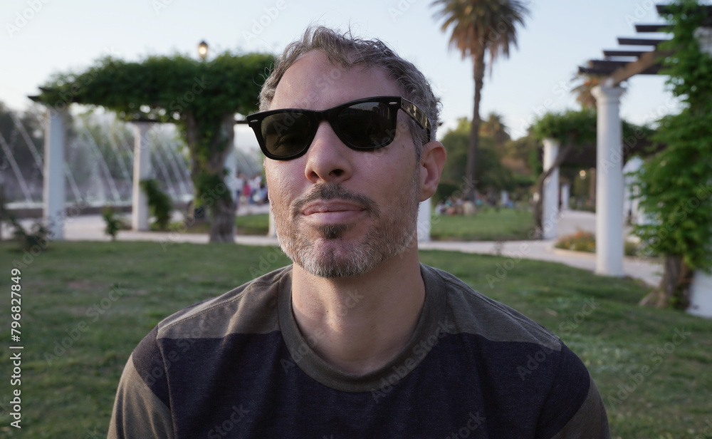 Portrait of a man in his 30s, wearing sunglasses sitting in the grass in the park at sunset. The white pergola columns in the background.
Portrait of a man in his 30s, wearing sunglasses sitting in the grass in the park at sunset. The white pergola columns in the background.