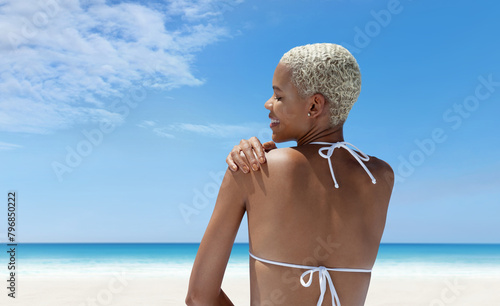 Portrait of a woman on the hot summer beach, enjoying a holiday by the sea. Seen from behind, with her hand on her shoulder, smiling towards the horizon of the ocean in front of her under a blue sky
