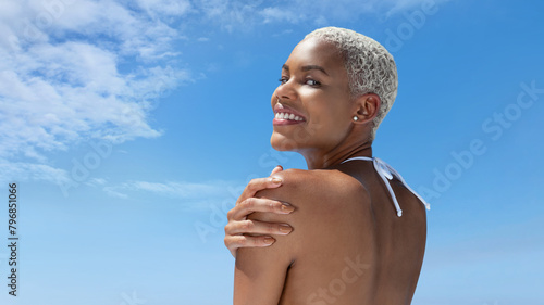 Portrait of a woman on the hot summer beach, enjoying a holiday by the sea. Seen from behind, with her hand on her shoulder, smiling towards the horizon of the ocean in front of her under a blue sky