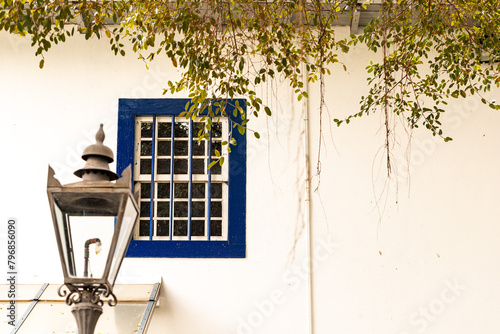 Poste de ferro, janela de madeira de cor azul, plantas no telhado e parede branca.  photo