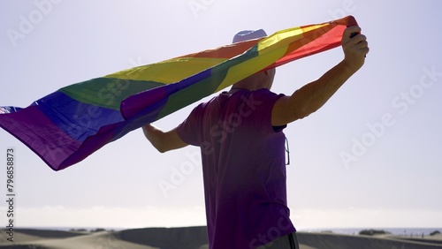 Gay hispanic man in purple t-shirt and tourist hat raises the rainbow flag on the sand dunes of Maspalomas. Gay tourism concepts photo