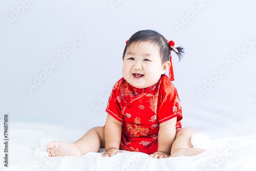 Chinese New Year concept, cute happy little girl in Chinese dress sitting in white living room. Cute Asian baby sitting in white living room, Chinese New Year concept.