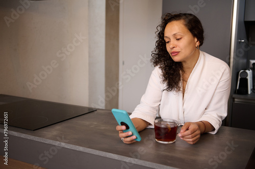 Happy young lady drinking tea and looking at smartphone, standing in modern minimalist home kitchen interior. Copy advertising space. Chat in morning, news and breakfast at home.