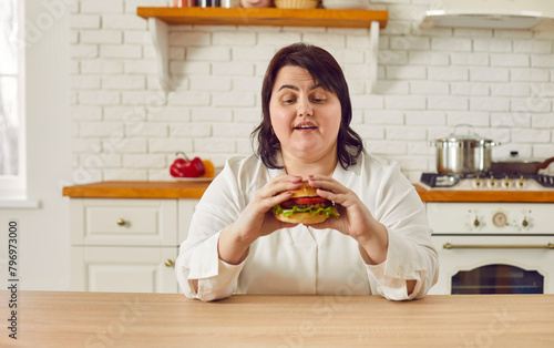 Overweight young woman eating hamburger while sitting at table in kitchen. Hungry plus size woman enjoying eating of delicious junk food. Unhealthy meal  overeating  extra calories