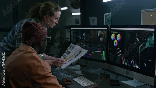 Medium side of Caucasian female postproduction studio producer and black male colorist looking at script and storyboard, discussing current project, scrolling through footage, pointing to frames