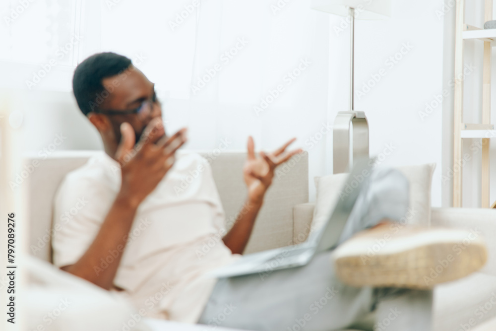 Smiling African American Freelancer Working on Laptop in Modern Home Office A young African American man sits on a comfortable sofa in his modern living room, working on his laptop with a smile on his Smiling African American Freelancer Working on Laptop in Modern Home Office A young African American man sits on a comfortable sofa in his modern living room, working on his laptop with a smile on his