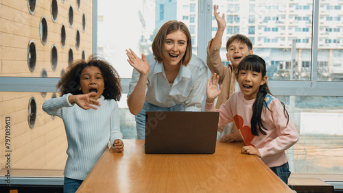 Happy caucasian teacher and multiethnic children waving hands to camera in casual uniform. Smart mentor and diverse student looking at laptop while greeting by moving hands or say good bye. Erudition. © Summit Art Creations