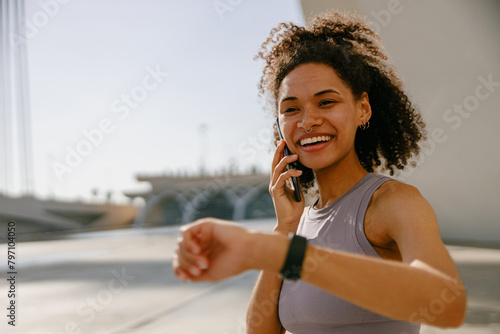 Smiling woman in sportswear looking on smartwatch after training outdoors while talking phone photo