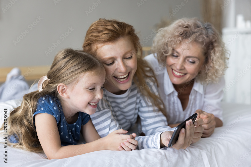 Close up happy little girl spend time together with young mother and grandmother lying together on bed, look at device screen, watching videos on internet, having fun, enjoy wireless tech, take selfie