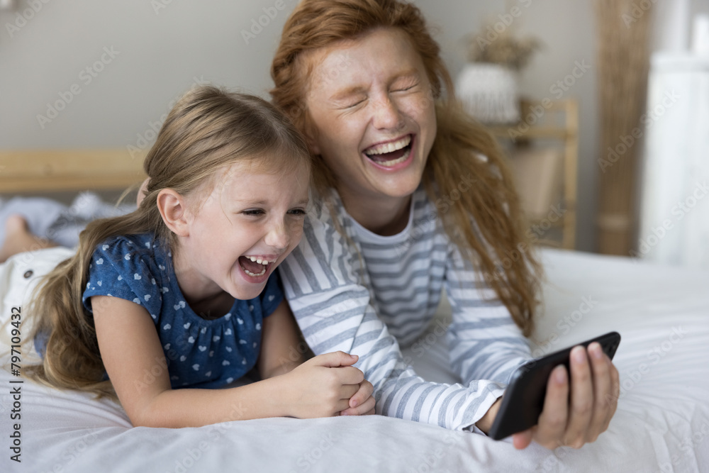 Joyful little cute girl relax in bed with mother on weekend at home, watch funny video or comedy movie, use smartphone online application, laughing. Happy family of two generation enjoy modern tech Joyful little cute girl relax in bed with mother on weekend at home, watch funny video or comedy movie, use smartphone online application, laughing. Happy family of two generation enjoy modern tech